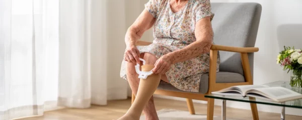 Femme âgée assise enfilant un bas de contention beige avec un accessoire dans son salon